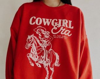 Op de afbeelding: Rood sweatshirt met een witte afbeelding van een vrouw die op een paard rijdt. De afbeelding zegt "COWGIRL Era" en "WE THE BABES WESTERN CO."