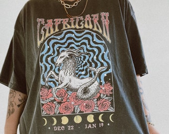 Könnte beinhalten: Ein überdimensioniertes, dunkelgraues T-Shirt mit einem Grafikdruck eines Steinbock-Sternzeichen mit einer Ziege und einem Fischschwanz, umgeben von Rosen und Monden. Der Text "CAPRICORN" ist in gelben und orangen Buchstaben über dem Grafikdruck gedruckt. Der Text "DEC 22 - JAN 19" ist unter dem Grafikdruck gedruckt.