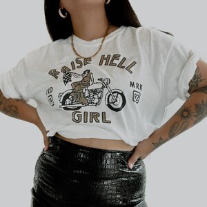 May include: White t-shirt with a graphic of a woman riding a motorcycle with the text "Raise Hell Girl" and "MRK 92". The woman is holding a flag that says "CRD 19".
