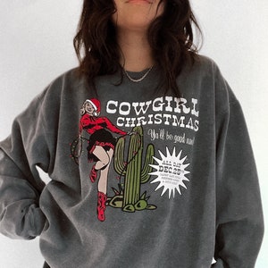 May include: Dark gray sweatshirt with a graphic that reads "COWGIRL CHRISTMAS" with a pin-up cowgirl in a Santa hat and red boots. The graphic includes a cactus and the text "Ya'll be good now!" and "DEC 25".