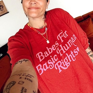 May include: A woman wearing a red t-shirt with the text "Babes For Basic Human Rights" in pink lettering. She has several tattoos on her arm, including a floral design, a crescent moon, and the words "Hold On Strong & Don't Let Go".