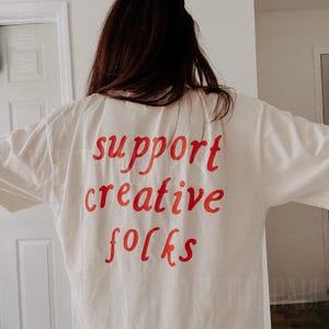 K&ouml;nnte beinhalten: Cremefarbenes T-Shirt mit dem roten Schriftzug "support creative folks" auf dem R&uuml;cken. Das Shirt besteht aus einem weichen Material und hat eine lockere Passform. Die Person, die das Shirt tr&auml;gt, hat lange braune Haare.