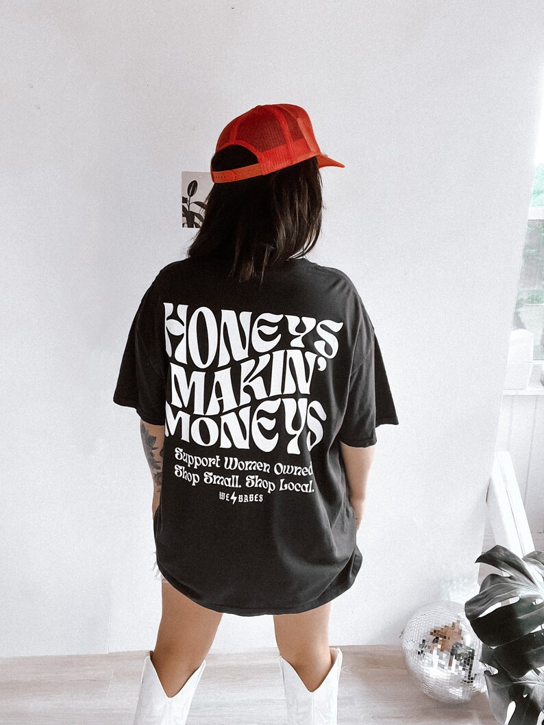 May include: Black t-shirt with white text that reads "Honeys Makin' Moneys" and "Support Women Owned Shop Small Shop Local" with a small lightning bolt and the words "We ⚡️ Babes".