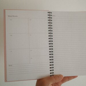 K&ouml;nnte beinhalten: Ge&ouml;ffneter Spiralplaner mit rosa Einband. Die linke Seite ist in Abschnitte f&uuml;r Woche und Monat unterteilt, mit Feldern f&uuml;r t&auml;gliche Eintr&auml;ge. Die rechte Seite hat liniertes Papier f&uuml;r Notizen.