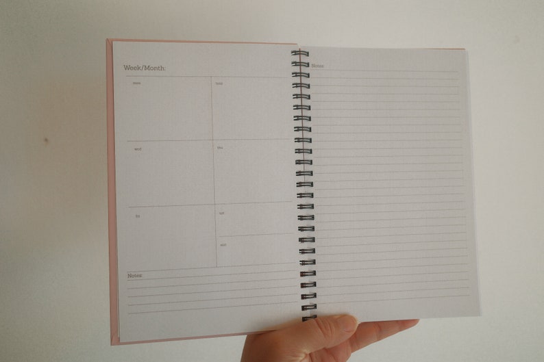 K&ouml;nnte beinhalten: Ge&ouml;ffneter Spiralplaner mit rosa Einband. Die linke Seite ist in Abschnitte f&uuml;r Woche und Monat unterteilt, mit Feldern f&uuml;r t&auml;gliche Eintr&auml;ge. Die rechte Seite hat liniertes Papier f&uuml;r Notizen.