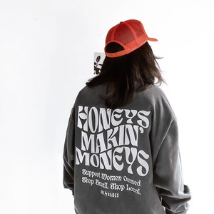 May include: A charcoal gray sweatshirt with the text "Honeys Makin Moneys" in white, stylized lettering. The text "Support Women Owned Shop Small Shop Local" is printed below in smaller white lettering. The sweatshirt has a relaxed fit and long sleeves. A red mesh baseball cap is worn on top of the head.