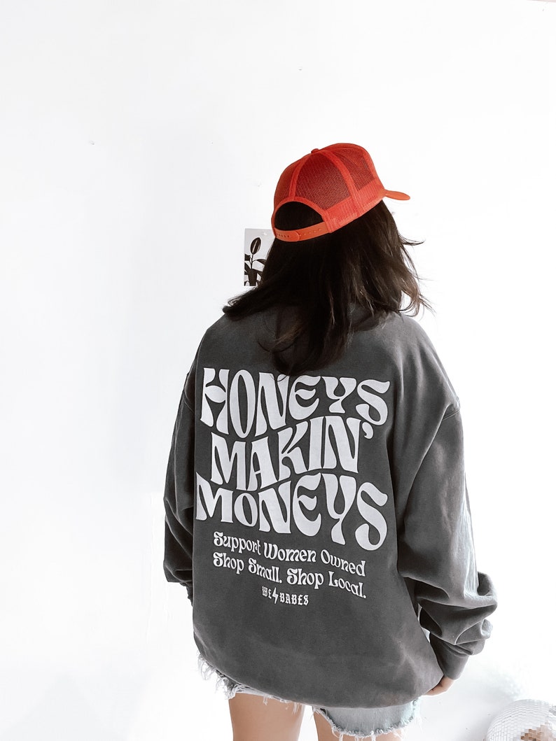 May include: A charcoal gray sweatshirt with the text "Honeys Makin Moneys" in white, stylized lettering. The text "Support Women Owned Shop Small Shop Local" is printed below in smaller white lettering. The sweatshirt has a relaxed fit and long sleeves. A red mesh baseball cap is worn on top of the head.
