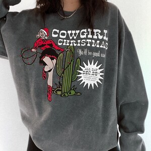 May include: Dark gray sweatshirt with a graphic that reads "COWGIRL CHRISTMAS" featuring a cowgirl in a Santa hat, cactus, and Christmas lights. The text "Y'all be good now" is also included.