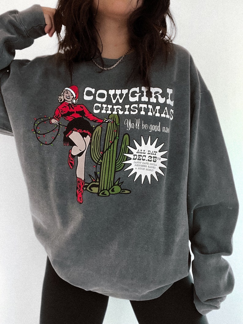 May include: Dark gray sweatshirt with a graphic that reads "COWGIRL CHRISTMAS" featuring a cowgirl in a Santa hat, cactus, and Christmas lights. The text "Y'all be good now" is also included.