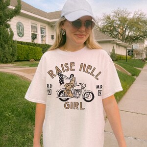 May include: White t-shirt with a graphic of a woman riding a motorcycle with the text "Raise Hell Girl" and "RD 19 MR 9" on either side of the motorcycle.