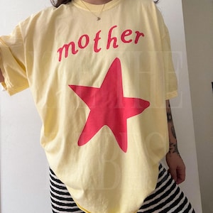 May include: A light yellow t-shirt with the word "mother" in red script above a large red star. The shirt is paired with black and white striped pants. The overall style is casual and comfortable.