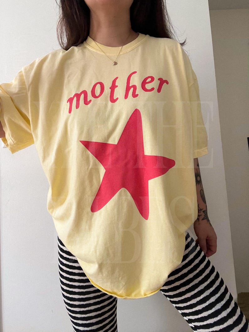 May include: A light yellow t-shirt with the word "mother" in red script above a large red star. The shirt is paired with black and white striped pants. The overall style is casual and comfortable.
