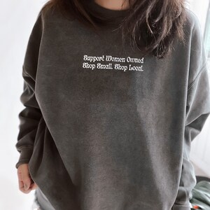 May include: A dark gray sweatshirt with the text "Support Women Owned Shop Small. Shop Local." in white lettering.
