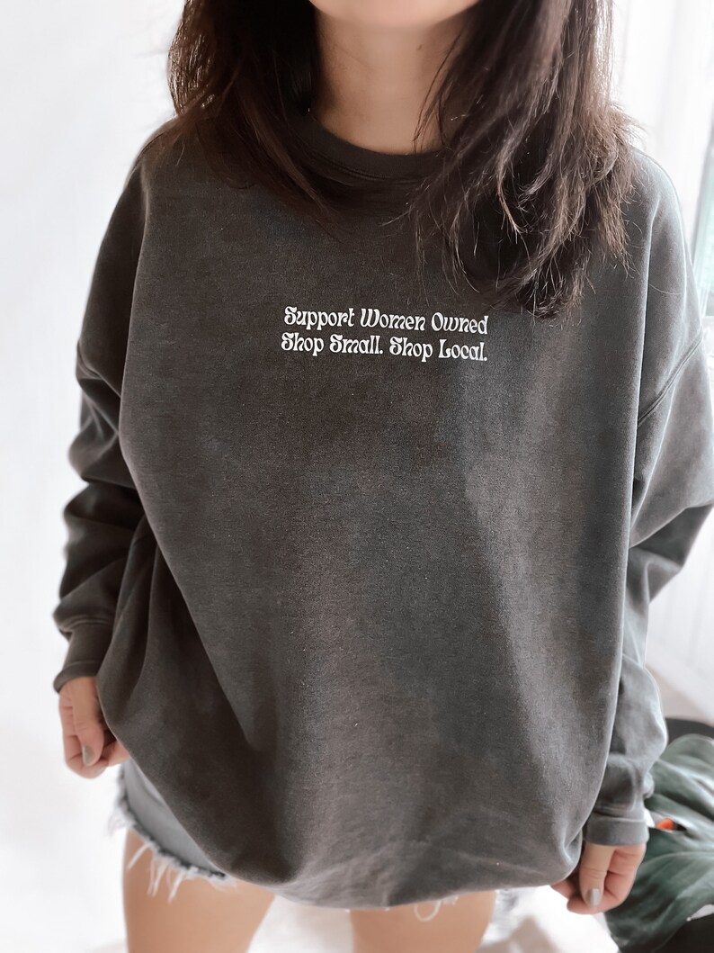 May include: A dark gray sweatshirt with the text "Support Women Owned Shop Small. Shop Local." in white lettering.