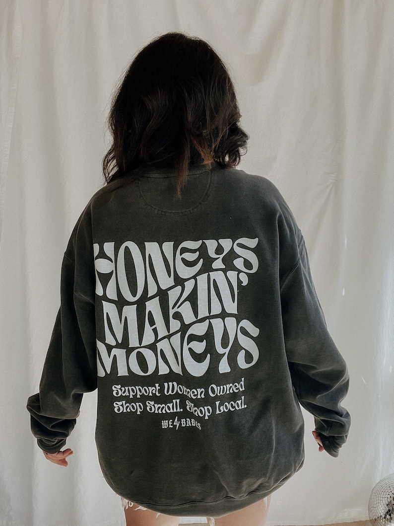May include: A dark gray sweatshirt with the text "Honeys Makin' Moneys" in white. The text is in a retro font style. Below the text is the phrase "Support Women Owned Shop Small Shop Local." in smaller white text. The text "WE 4 BABIS" is also in white text.