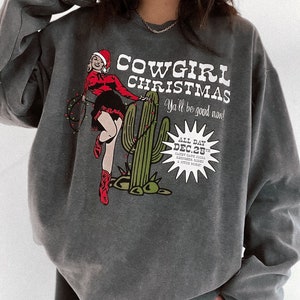 May include: Dark gray sweatshirt with a graphic that reads "Cowgirl Christmas" featuring a cowgirl in a Santa hat and cactus with Christmas lights. The text "Ya'll be good now!" is also included.