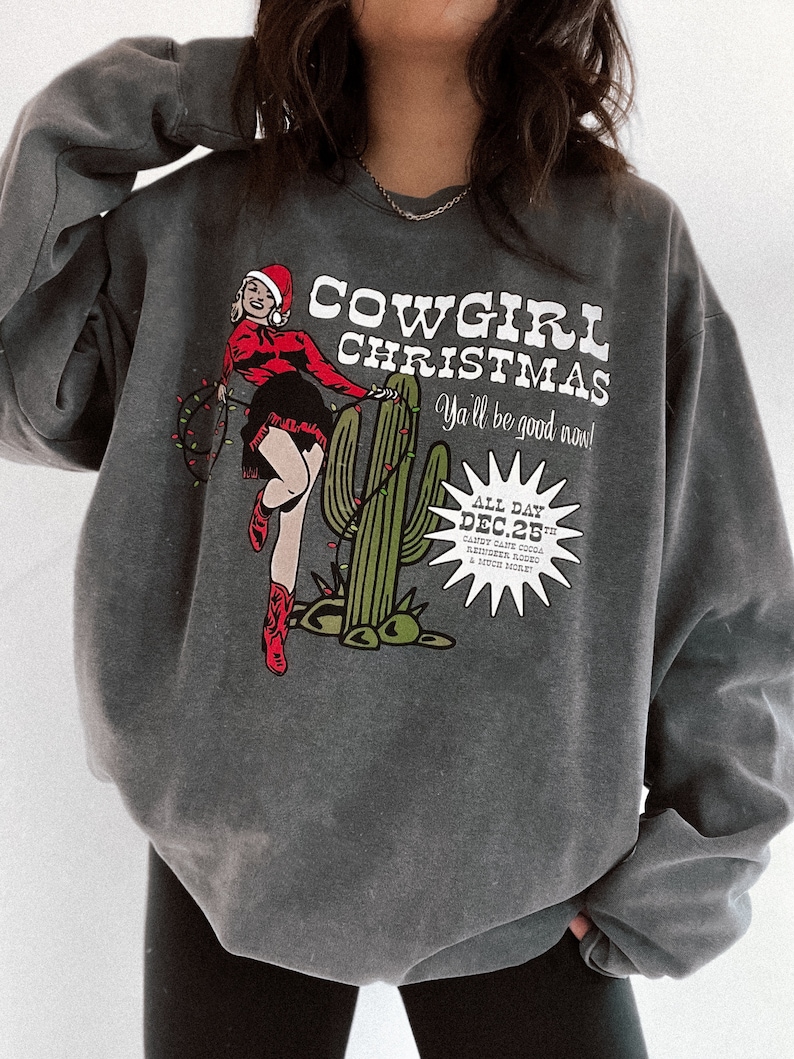 May include: Dark gray sweatshirt with a graphic that reads "Cowgirl Christmas" featuring a cowgirl in a Santa hat and cactus with Christmas lights. The text "Ya'll be good now!" is also included.