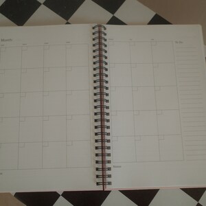 K&ouml;nnte beinhalten: Ge&ouml;ffneter Spiralplaner mit monatlichem Kalenderlayout. Die Seiten sind wei&szlig; mit einem Raster von K&auml;stchen f&uuml;r jeden Tag und einem Abschnitt f&uuml;r Notizen und To-Do-Listen. Die Bindung ist schwarz und der Einband hellrosa.