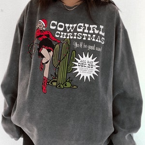 May include: Dark gray sweatshirt with a graphic that reads "COWGIRL CHRISTMAS" featuring a cowgirl in a Santa hat, cactus, and Christmas lights. The text "Ya'll be good now!" is also included. The date "DEC. 25th" is also visible.