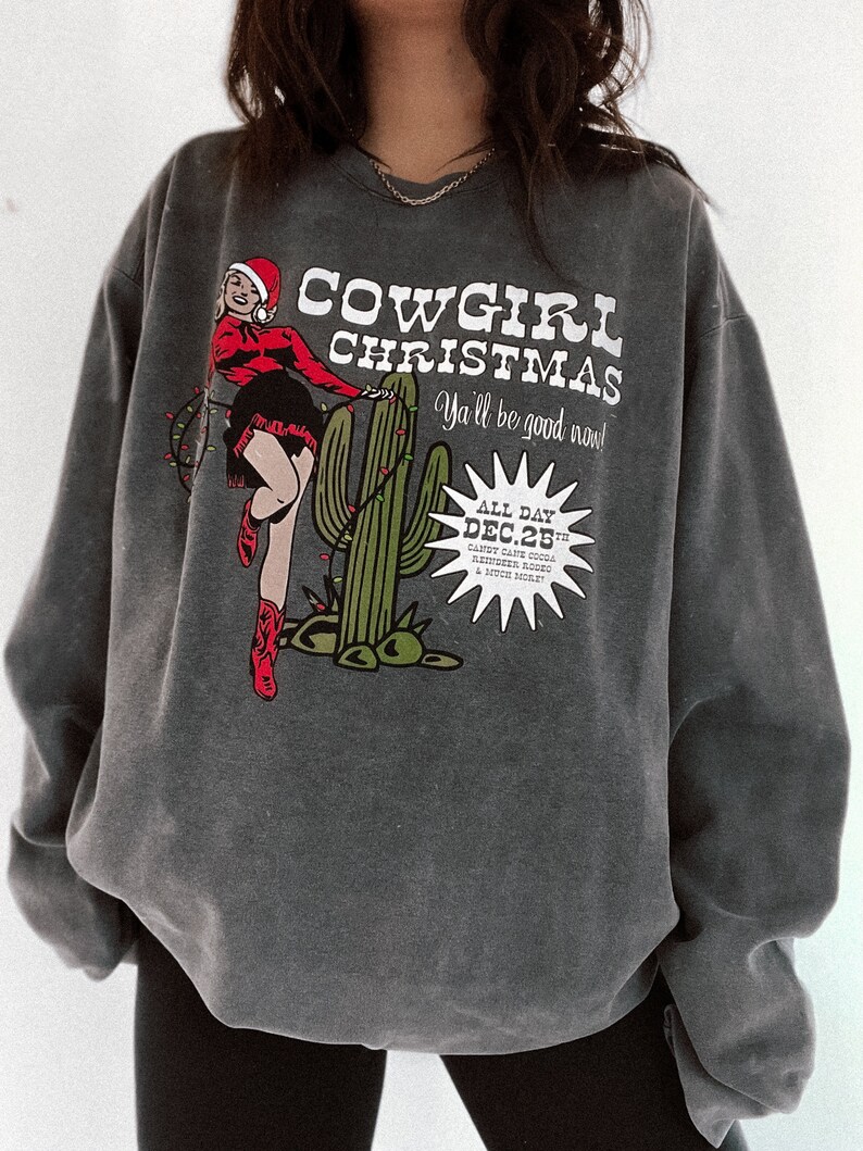 May include: Dark gray sweatshirt with a graphic that reads "COWGIRL CHRISTMAS" featuring a cowgirl in a Santa hat, cactus, and Christmas lights. The text "Ya'll be good now!" is also included. The date "DEC. 25th" is also visible.