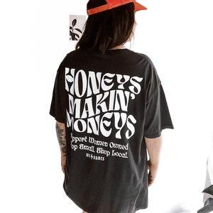 May include: Black oversized t-shirt with white text that reads "Honeys Makin' Moneys" and "Support Women Owned. Shop Small. Shop Local. We4Babes". The model is wearing a red mesh baseball cap and white cowboy boots.