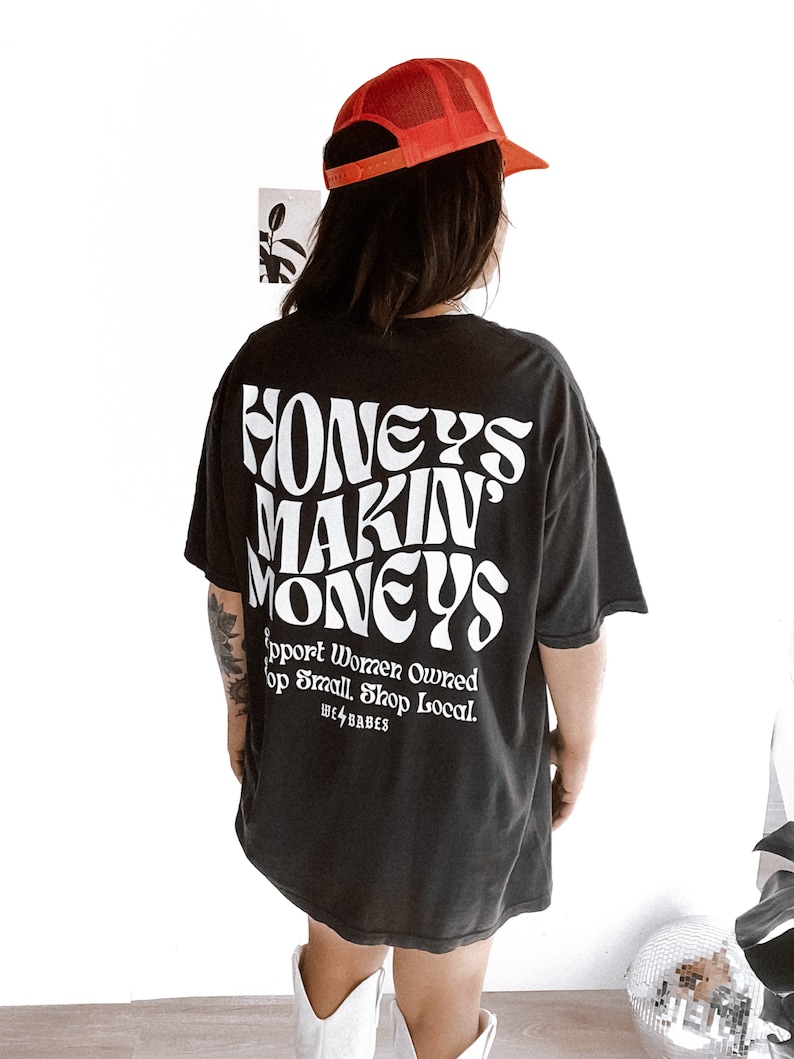 May include: Black oversized t-shirt with white text that reads "Honeys Makin' Moneys" and "Support Women Owned. Shop Small. Shop Local. We4Babes". The model is wearing a red mesh baseball cap and white cowboy boots.