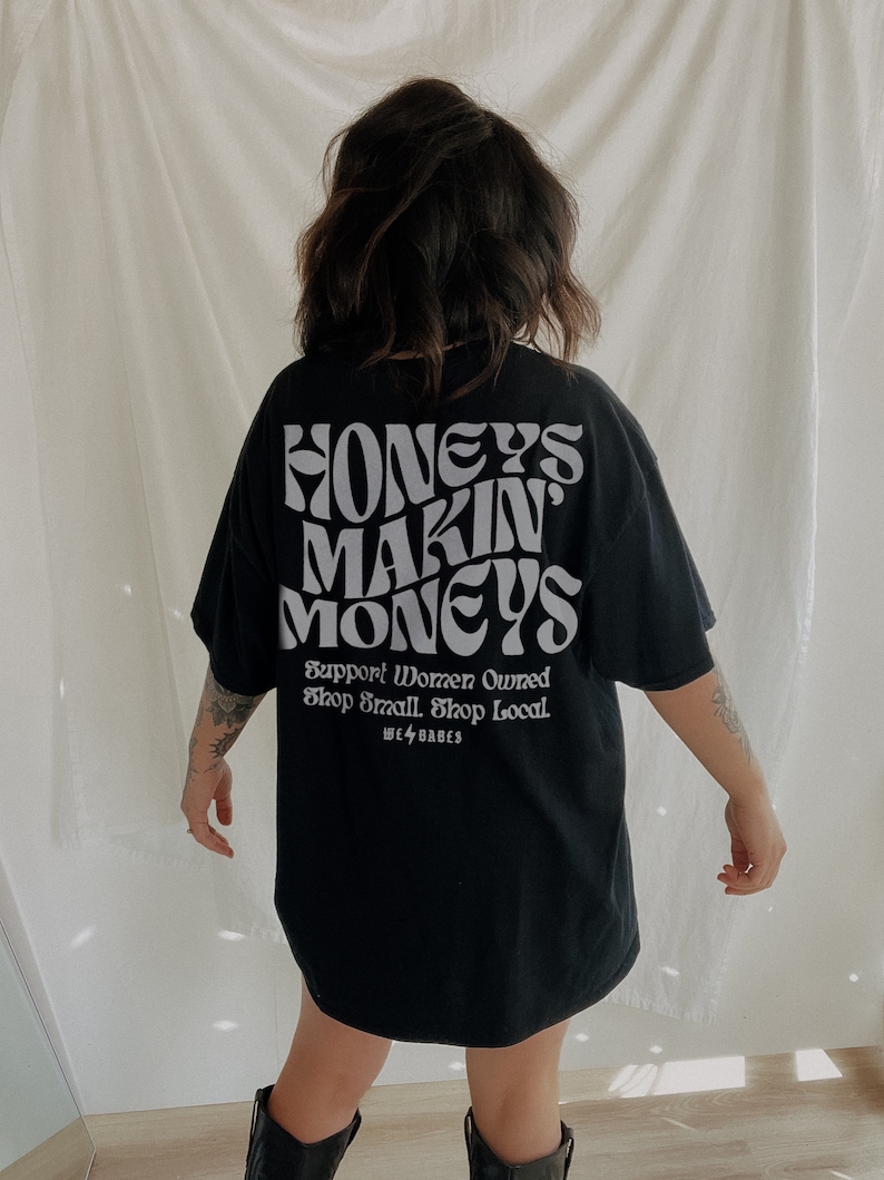 May include: Black t-shirt with the text "Honeys Makin' Moneys" in white, with the slogan "Support Women Owned Shop Small Shop Local" and the logo "We<3Babes" underneath.