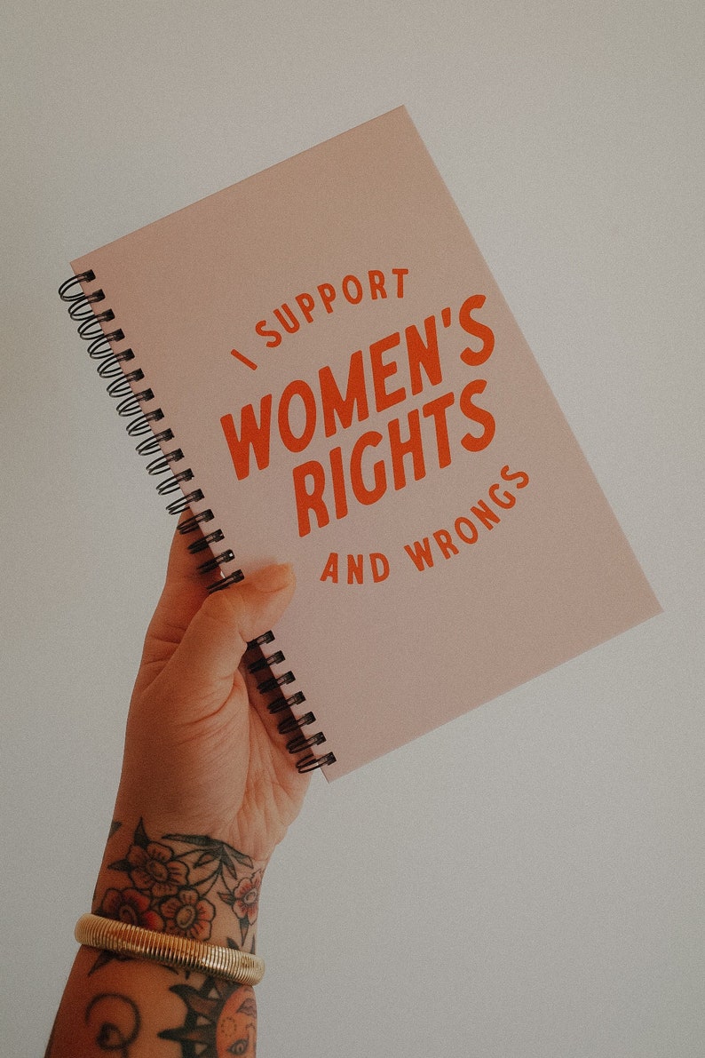 K&ouml;nnte beinhalten: Ein rosa Spiralnotizbuch mit dem Text "I SUPPORT WOMEN'S RIGHTS AND WRONGS" in Orange. Das Notizbuch wird in einer Hand mit einem goldenen Armband und floralen T&auml;towierungen gehalten.