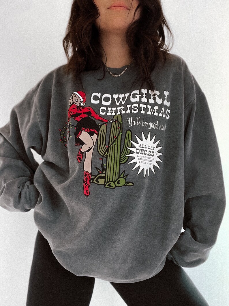 May include: Dark gray sweatshirt with a graphic that reads "COWGIRL CHRISTMAS" with a pin-up cowgirl in a Santa hat and red boots. The graphic includes a cactus and the text "Ya'll be good now!" and "DEC 25".