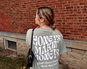 May include: A sage green t-shirt with the text "Honeys Makin Money" in a retro font. The shirt also has the text "Support Women Owned Small. Shop Local. We4Babes" printed on the back.