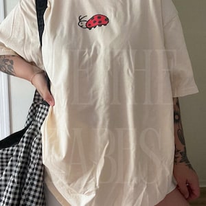 K&ouml;nnte beinhalten: Cremefarbenes T-Shirt mit einem roten und schwarzen Marienk&auml;fer-Motiv auf der Vorderseite. Das Shirt hat kurze &Auml;rmel und eine lockere Passform. Eine schwarz-wei&szlig; karierte Tasche ist im Hintergrund zu sehen.