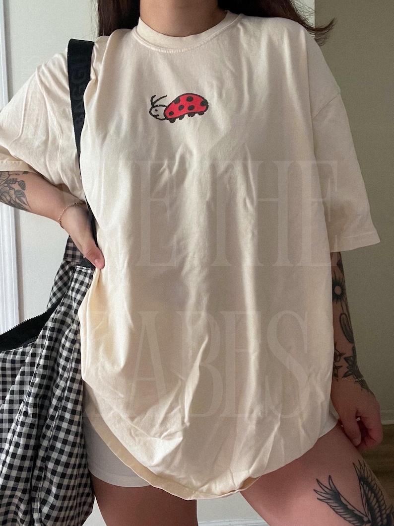 K&ouml;nnte beinhalten: Cremefarbenes T-Shirt mit einem roten und schwarzen Marienk&auml;fer-Motiv auf der Vorderseite. Das Shirt hat kurze &Auml;rmel und eine lockere Passform. Eine schwarz-wei&szlig; karierte Tasche ist im Hintergrund zu sehen.
