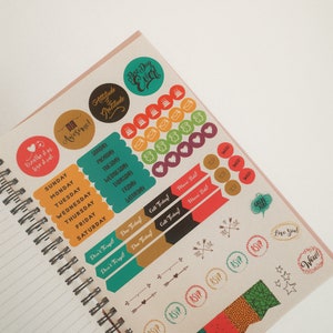 K&ouml;nnte beinhalten: Offenes Notizbuch mit Spiralbindung und bunten Aufklebern. Aufkleber mit S&auml;tzen wie "Best Day Ever", "Attitude of Gratitude" und Tagesetiketten. Andere Aufkleber haben RSVP- und Pfeildesigns.