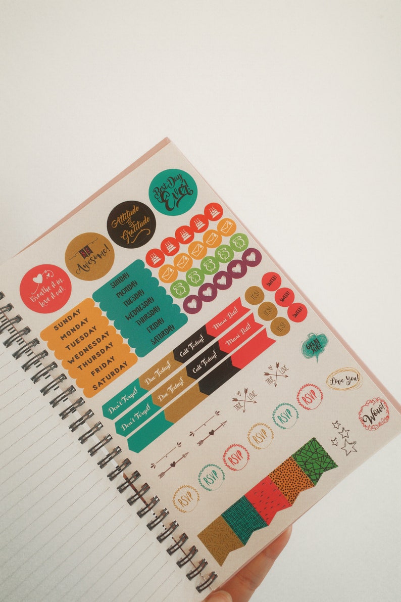 K&ouml;nnte beinhalten: Offenes Notizbuch mit Spiralbindung und bunten Aufklebern. Aufkleber mit S&auml;tzen wie "Best Day Ever", "Attitude of Gratitude" und Tagesetiketten. Andere Aufkleber haben RSVP- und Pfeildesigns.