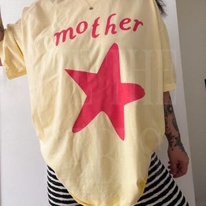 May include: A light yellow t-shirt with the word "mother" in red script above a large red star. The shirt is paired with black and white striped leggings. The overall style is casual and relaxed.