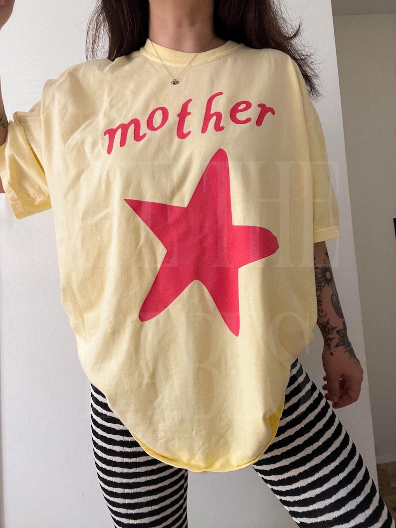 May include: A light yellow t-shirt with the word "mother" in red script above a large red star. The shirt is paired with black and white striped leggings. The overall style is casual and relaxed.