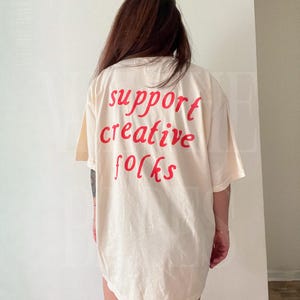 K&ouml;nnte beinhalten: Cremefarbenes &uuml;bergro&szlig;es T-Shirt mit dem roten Aufdruck "support creative folks" auf dem R&uuml;cken. Das Shirt hat kurze &Auml;rmel und eine lockere Passform, geeignet f&uuml;r Freizeitkleidung.