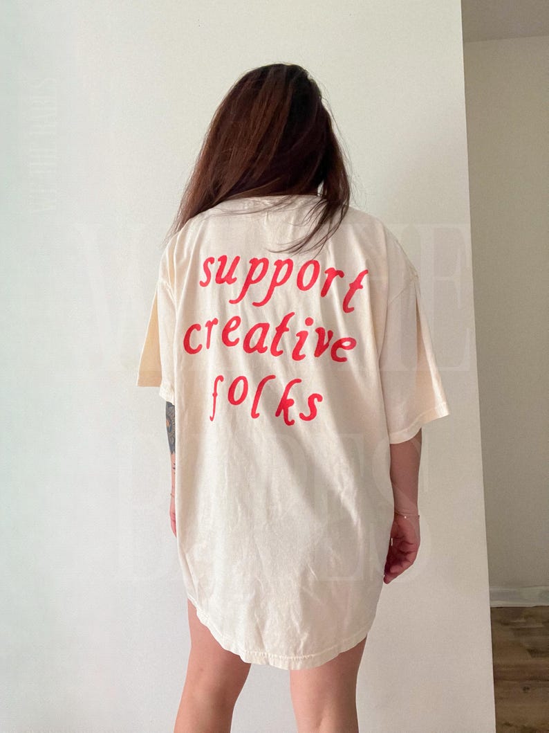 K&ouml;nnte beinhalten: Cremefarbenes &uuml;bergro&szlig;es T-Shirt mit dem roten Aufdruck "support creative folks" auf dem R&uuml;cken. Das Shirt hat kurze &Auml;rmel und eine lockere Passform, geeignet f&uuml;r Freizeitkleidung.