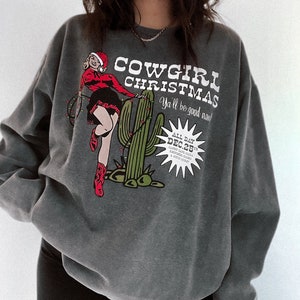 May include: Dark gray sweatshirt with a graphic that reads "COWGIRL CHRISTMAS" with a cartoon cowgirl in a Santa hat and red boots. A green cactus is also featured in the design. The text "Ya'll be good now!" is also included.