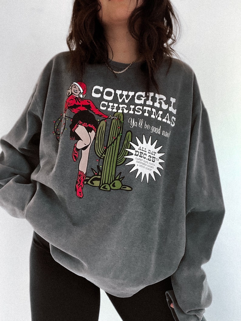 May include: Dark gray sweatshirt with a graphic that reads "COWGIRL CHRISTMAS" with a cartoon cowgirl in a Santa hat and red boots. A green cactus is also featured in the design. The text "Ya'll be good now!" is also included.