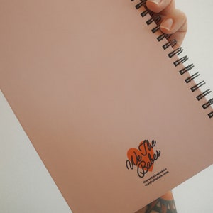 K&ouml;nnte beinhalten: Ein rosafarbenes Notizbuch mit Spiralbindung. Das Notizbuch ist ge&ouml;ffnet und zeigt leere Seiten. In der unteren rechten Ecke befindet sich ein Logo mit dem Text "We The Babes" in einem orangefarbenen Herz und eine Website-Adresse.