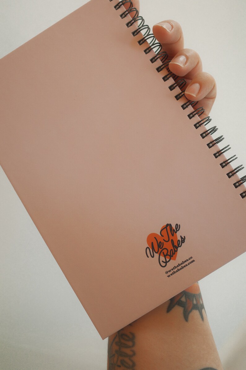 K&ouml;nnte beinhalten: Ein rosafarbenes Notizbuch mit Spiralbindung. Das Notizbuch ist ge&ouml;ffnet und zeigt leere Seiten. In der unteren rechten Ecke befindet sich ein Logo mit dem Text "We The Babes" in einem orangefarbenen Herz und eine Website-Adresse.