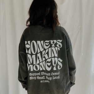 May include: A dark gray sweatshirt with the text "HONEY'S MAKIN' MONEYS" in white. The text is in a retro font style. Below the text is the phrase "Support Women Owned Shop Small. Shop Local." in white. The text "WE 4 BABES" is also in white.
