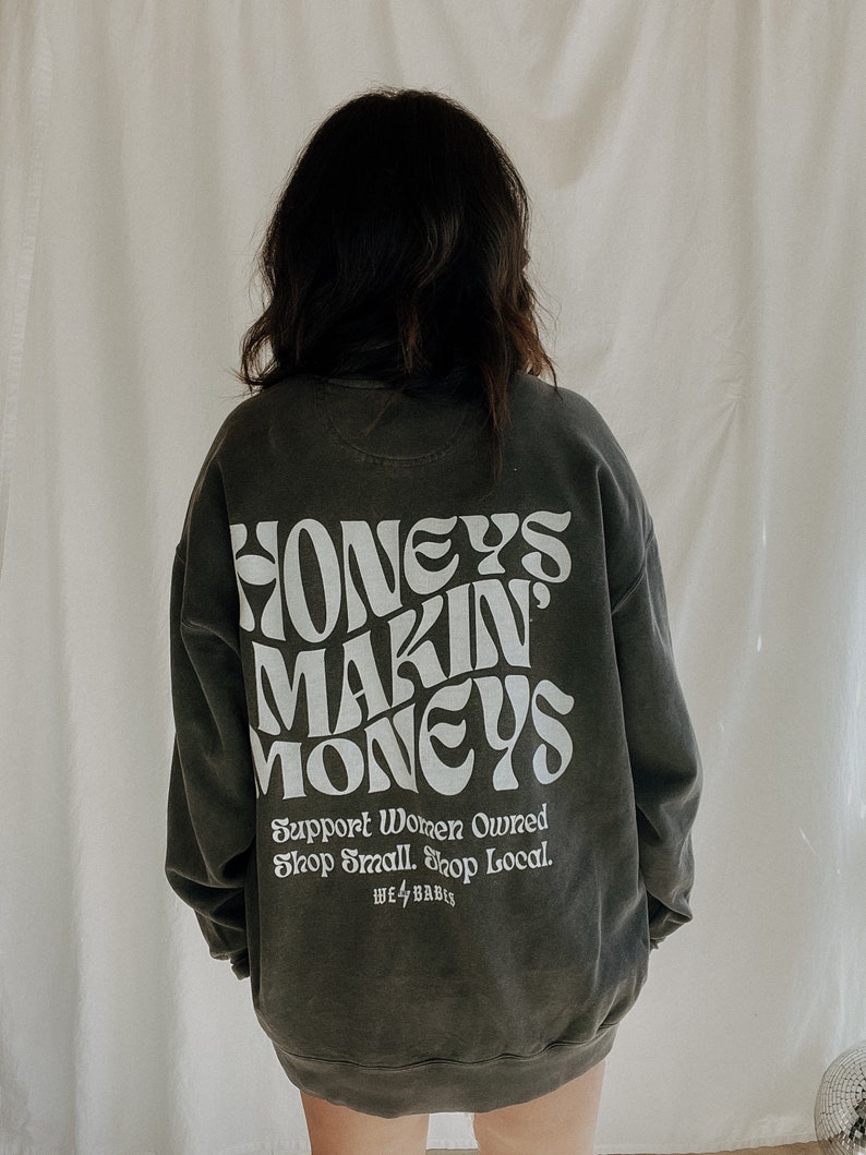 May include: A dark gray sweatshirt with the text "HONEY'S MAKIN' MONEYS" in white. The text is in a retro font style. Below the text is the phrase "Support Women Owned Shop Small. Shop Local." in white. The text "WE 4 BABES" is also in white.