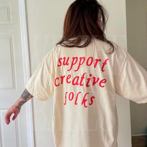 K&ouml;nnte beinhalten: Cremefarbenes &uuml;bergro&szlig;es T-Shirt mit dem roten Aufdruck "support creative folks". Das Shirt hat kurze &Auml;rmel und eine lockere Passform. Der Text ist im handschriftlichen Stil gehalten.