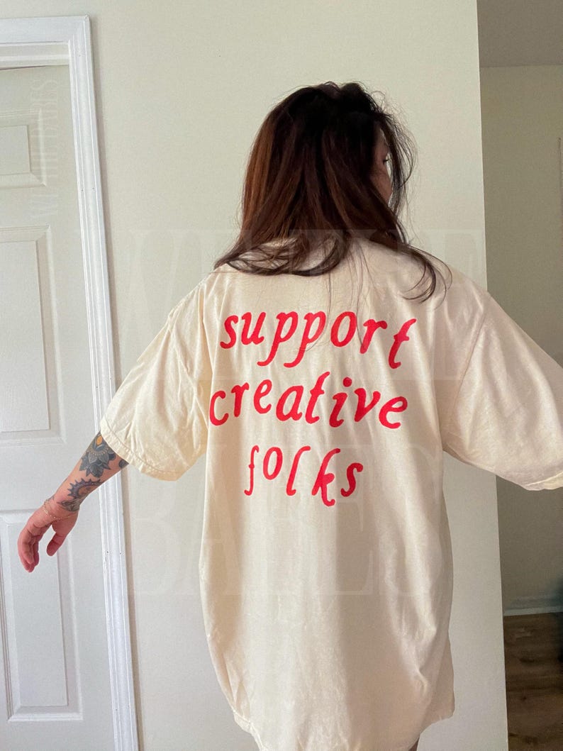 K&ouml;nnte beinhalten: Cremefarbenes &uuml;bergro&szlig;es T-Shirt mit dem roten Aufdruck "support creative folks". Das Shirt hat kurze &Auml;rmel und eine lockere Passform. Der Text ist im handschriftlichen Stil gehalten.