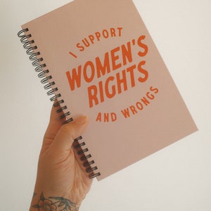 K&ouml;nnte beinhalten: Ein rosa Spiralnotizbuch mit dem Text "I SUPPORT WOMEN'S RIGHTS AND WRONGS" in orangefarbenen Buchstaben. Das Notizbuch wird in einer Hand gehalten, mit einem goldenen Armband und einem floralen Tattoo.