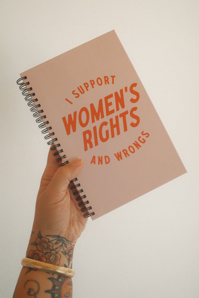 K&ouml;nnte beinhalten: Ein rosa Spiralnotizbuch mit dem Text "I SUPPORT WOMEN'S RIGHTS AND WRONGS" in orangefarbenen Buchstaben. Das Notizbuch wird in einer Hand gehalten, mit einem goldenen Armband und einem floralen Tattoo.
