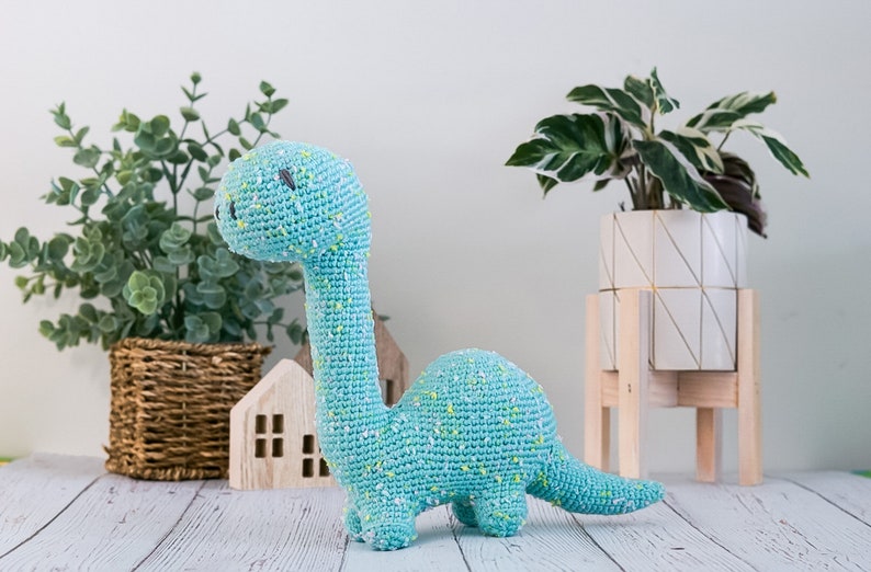 Puede incluir: Un juguete de dinosaurio de crochet azul claro con motas amarillas. El dinosaurio est&aacute; de pie sobre una superficie de madera blanca con una cesta de mimbre y una peque&ntilde;a casa de madera detr&aacute;s.