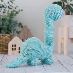 Puede incluir: Un juguete de dinosaurio de crochet azul claro con detalles blancos y amarillos. El dinosaurio est&aacute; de pie sobre una superficie de madera blanca.
